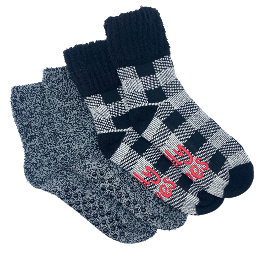 One black slipper sock with a black and white plaid pattern featuring a solid black cuff. Also features a solid black heel and toe patch. The bottom contains a rubber gripper in the shape of text Jolly Vibes. 